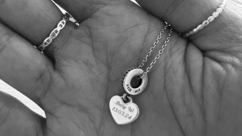 A black and white image of the palm of a hand holding a necklace with the words Baby W 13.03.24