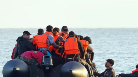 A group of migrants, some wearing life jackets are in an inflatable boat, with two more people in the water who are trying to climb aboard.