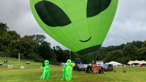 Green alien hot air balloon, with two green alien costumes in front of it 