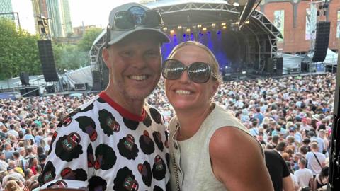 Kaela and Jonathan Rigg stand together at a concert. She is wearing sunglasses and he is wearing a bucket hat. There is a large crowd and a stage in the background. 