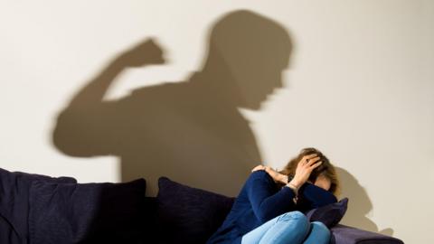 A shadow of a man with a clenched fist as a woman cowers in the corner of a room, on a sofa. 