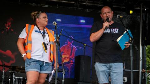 Ria and Luke Simons pictured on stage. Ria Simons is wearing an orange high viz jacket with a white top and denim shorts. Luke Simons has a black t-shirt with the words "Woo Fest" written on the front and is wearing blue shorts.