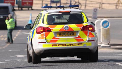 A police car parked on a road leading to a beach. There is a man wearing a hi-viz jacket standing to the left of the car.