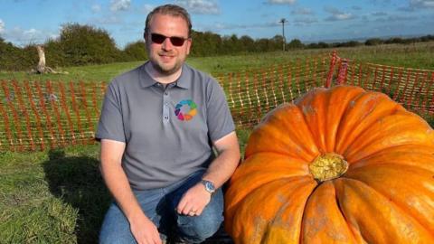 Ben Rayner is sitting on the grass next to his huge pumpkin. He is leaning against it. He is wearing a grey shirt and sunglasses.