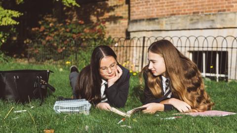 Two teenage schoolgirls in uniform lie on the grass reading and studying outdoors after class - stock image