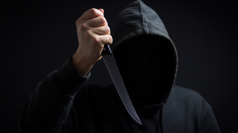 A man is wearing a black hooded top with his face hidden and is holding a knife