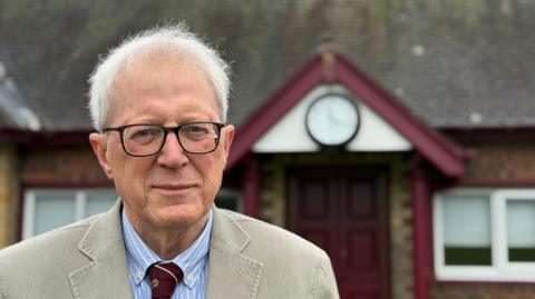 A elderly man stood in from of a maroon painted doorway that has an analogue clock above it. The man has grey thinning hair and glasses. He is wearing a cream coloured blazer with a striped canvas shirt and a maroon tie.