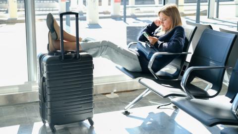 A young woman with a suitcase waiting for her flight at the airport, looking at her smartphone for a delayed departure