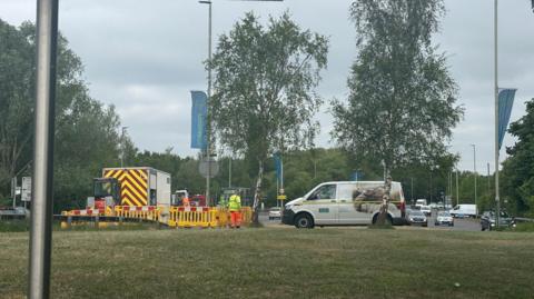 A team of two men from Severn Trent, wearing hi-vis clothes, stand near parked vans on a roadside grass verge near a busy road. 