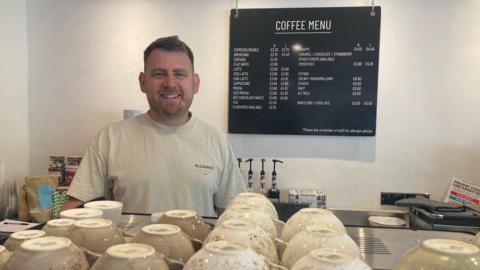 Bradley Buechel wears a grey t-shirt and looks into the camera from behind his professional coffee machine. A price list is behind him on the wall and there are coffee cups lined up on the top of the machine.