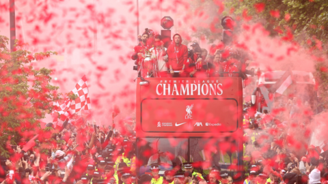 Image shows Liverpool FC's victory parade bus moving through Liverpool. Players stand on the open top deck as thousands of fans surround the vehicle. Confetti and smoke can be seen hanging in the air. 