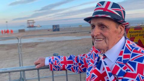 An elderly man pictured in front of a beach wearing a suit covered in Union flags and wearing a hat with the same pattern on it.