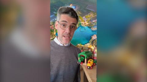 A man with glasses on is staring at the camera, the green screen back drop is an artist impression of Universal UK's site. There are wooden toy trains next to him. 