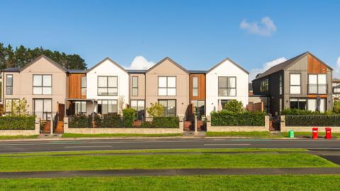 A row of modern render-fronted houses. There is a lawned area in the foreground. The sky is blue.