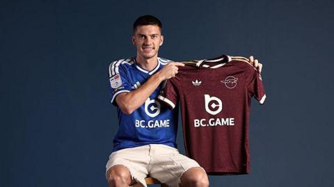 Admir Bristric, sat down while wearing a blue Leicester City shirt and white shorts, holding up a maroon away Leicester City shirt upwards with both hands to his left-hand side