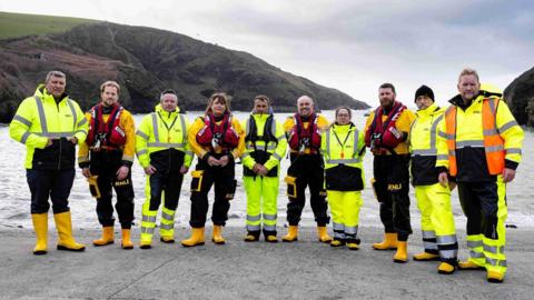Ten of the Port Isaac RNLI team are standing in a line in the harbour wearing their hi-vis uniforms and yellow wellies. The sea and a steep headland are behind them on this grey day.