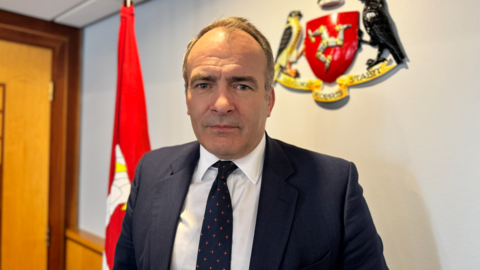 Alfred Cannan is wearing a dark blue suit, white shirt and blue tie with small red spots. He is clean shaven with dark receding hair. He is standing in an office with a large Manx flag to the left and an Isle of Man crest on the wall to the right.