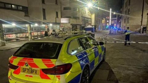 A police car parked in front of police tape on a dark street in edinburgh with police officers far in the background.