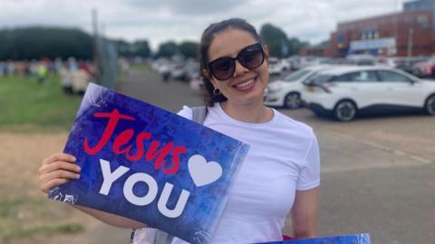 A woman with brown hair and sunglasses smiles at the camera, She is wearing a white t-shirt, and is holding a sign which reads 'Jesus (heart shape) YOU'. There are a number of parked cars behind the woman, as well as a large patch of grass, several trees, and a crowd of people. 