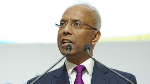 Mayor of Tower Hamlets Lutfur Rahman speaking at after he was elected. He is standing against a white background and is wearing a dark suit, while shite and purple tie. There are two microphones in front of him. 