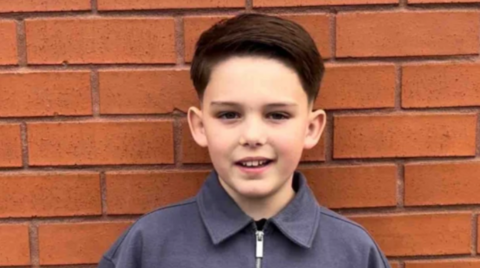 A headshot image of a young boy in a grey short, in front of a brick wall