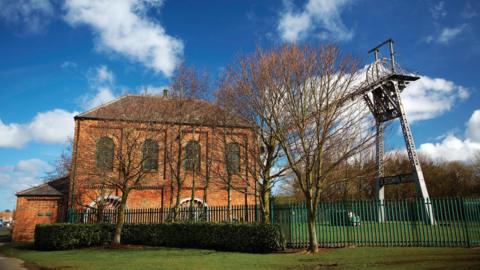 Washington F-Pit Museum. The red stone building with large green-framed arch windows is located behind a green, metal fence and a number of leafless trees. The large metal pit head stands to the right of it. A long section of the machinery leads down to the building.