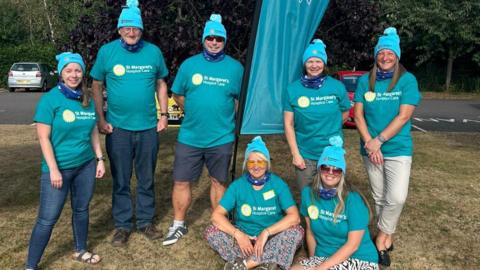 A group of seven people all dressed in blue T-shirts is standing next to each other on grass. They are wearing blue hats and are all smiling at the camera.