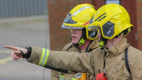 Two male firefighters dressed in beige uniform jackets with yellow fluorescent strips on arms and chests and yellow helmets with chin-straps. , one pointing at an object. The man on the left is pointing towards something unseen. They are standing outside, with a red brick building  and corrugated metal structure out-of-focus in the background.