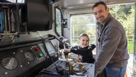 Cillian Gallagher and his father are in the locomotive playing with the buttons. They are both smiling at the camera.