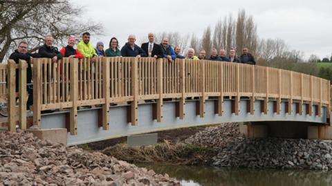 People lined up on a wooden bridge crossing a river. There are 16 people on the bridge with some of them leaning on the fence. 