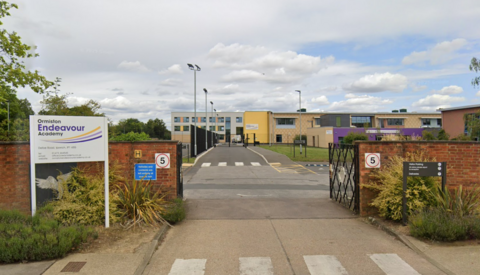 The entrance of Ormiston Endeavour Academy in Ipswich. The school premises is surrounded by a red brick wall. School buildings can be seen in the distance. A white welcome sign details the school name.