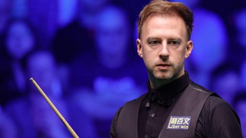 Judd Trump in black shirt and waistcoat carrying a snooker cue