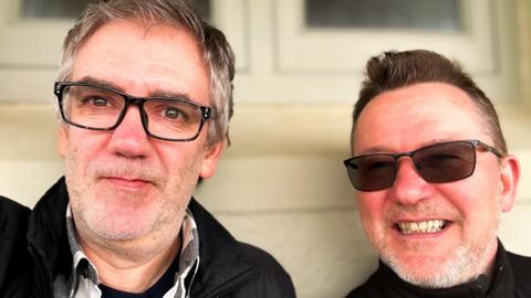 Stuart and Carl pose for a selfie. They both smile and are wearing glasses.