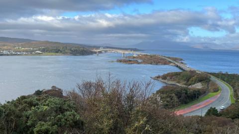 A grey road and bridge over a blue sea with some brown and green trees in the foreground, as white, fluffy clouds linger in the sky. 