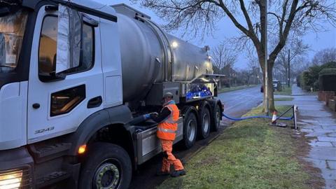A man dressed in a high-vis suit stands near a tanker lorry which appears to be plugged into a water supply. It is early morning light.