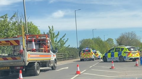 Part of the A10 where a road closure is in place. At the scene of the incident there are a number of marked police cars parked on the road and a highway maintenance van.