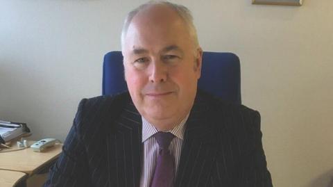 Malcolm Burr - a man with grey hair pictured sitting at his desk smiling wearing a pinstripe suit and purple tie