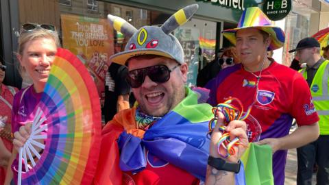 People dressed in rainbow colours, smiling on a hot summer's day. A man in the foreground is smiling, wearing black sunglasses. There are shops behind him.