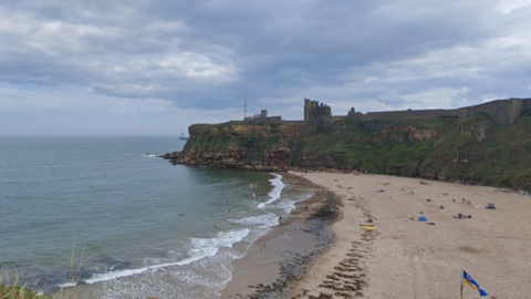 A zoomed out view of a curved stretch of beach with numerous people on the sand. The sea is to the left with large amounts of seaweed. Cliffs can be seen in the background with the ruins of Tynemouth Priory visible.