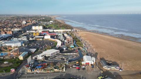 A wide aerial view of Skegness showing a large sandy beach stretching along the coastline beside calm blue-grey water. Next to the beach is a busy seafront area with amusement rides, go‑kart tracks, and various buildings, including arcades and attractions. Further inland, rows of houses and streets extend into the distance. The scene is bright and clear, with the coastline curving away towards the horizon.