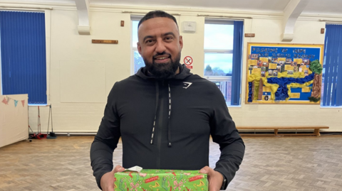 A man in a black jacket smiles at the camera as he hold a Christmas present wrapped in green and red wrapping paper. he stands in a school hall with blue blinds and a blue art display behind him.