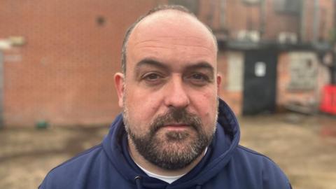 McCusker is looking directly at the camera. He has brown/grey facial hair and thin grey hair. He is wearing a navy hoodie.