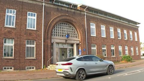 The front of Torquay police station. A silver car is driving along the road outside the red brick building which has several sash windows. A mini blue street light with the Devon and Cornwall Police emblem on it is above the front door.
