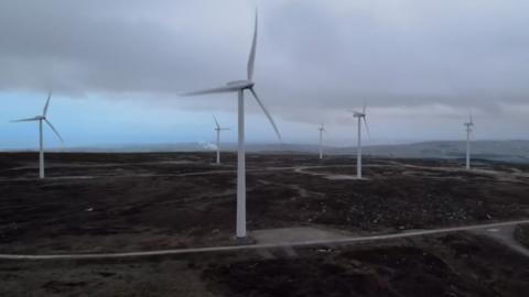 Six wind turbines on a dark brown moor with a large amount of cloud cover in the sky
