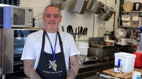 A man wearing a white t-shirt and a black apron stands in kitchen. Kitchen surfaces and equipment can be seen around him.