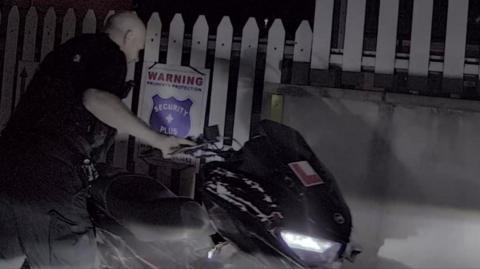 A bald police officer dressed in black uniform holds onto the handlebars of a black damaged moped while looking down at it. The headlight is on as it is dark. Behind the moped is a white fence with a warning sign on it.