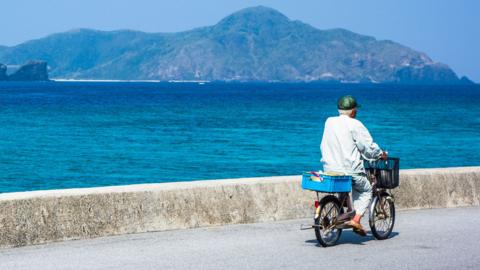A man with grey hair wearing a green cap and a light blue shirt riding a bike along a concrete path. He is next 