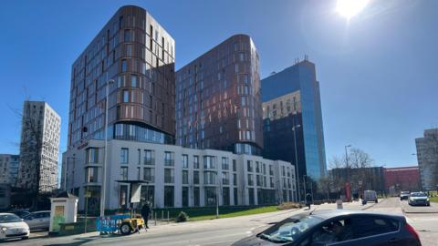Large tower blocks in Coventry city centre. The first two in the centre of the image are curved buildings in brown metallic colour. The building on their right is angular and reflective. The street below it seems empty with a handful of parked cars