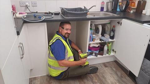 Man wearing high-vis, goggles and holding a drill sits underneath a kitchen sink looking at the camera