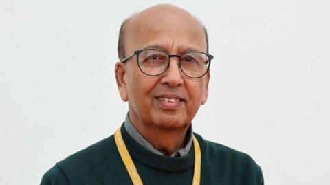 Prof Ahmed has heavily receding dark hair and is wearing black round spectacles on his face, a dark-green jumper over a sage-coloured shirt.